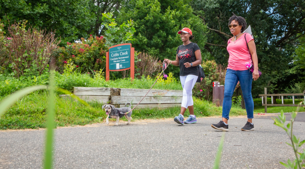 Two Black women walking a small dog on a leash along the Cobbs Creek Trail on the Circuit Trails