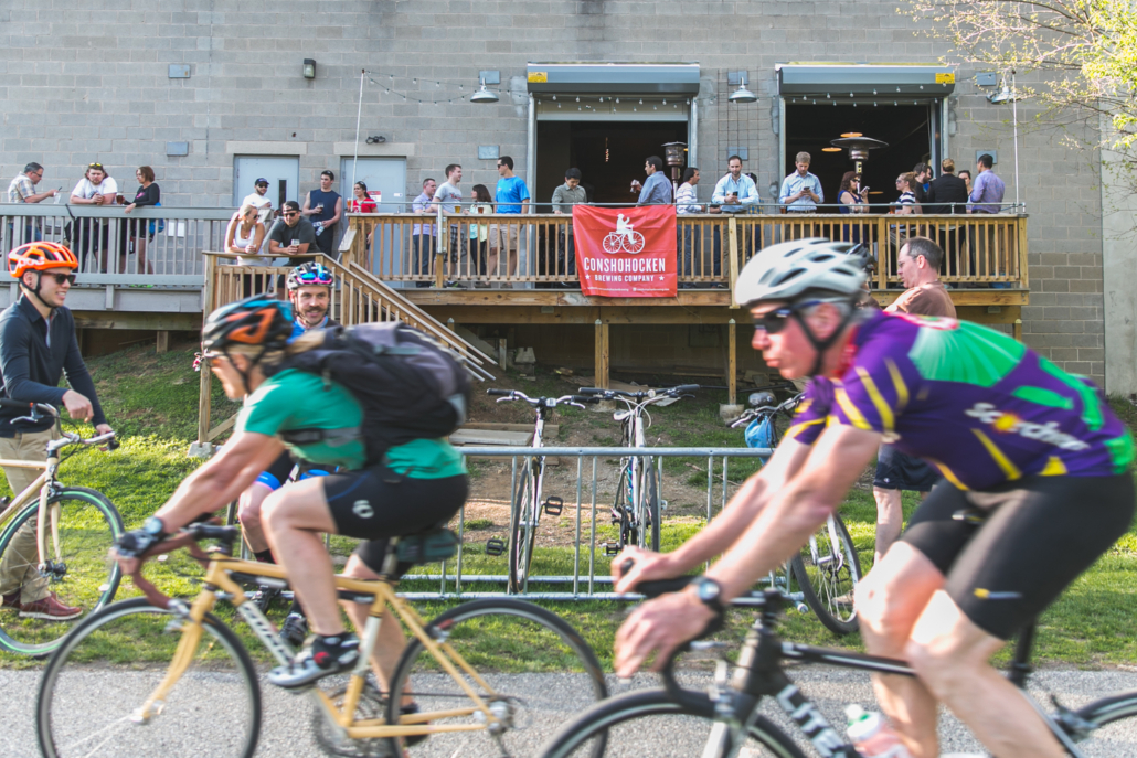 two men on bikes riding on the Schuylkill River Trail in front of people enjoying beverages on the porch of the Conshohocken Brewing Company Tap Room in Conshohocken