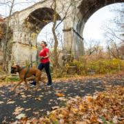 Woman in a red shirt walking a dog on a trail that has leaves on the ground