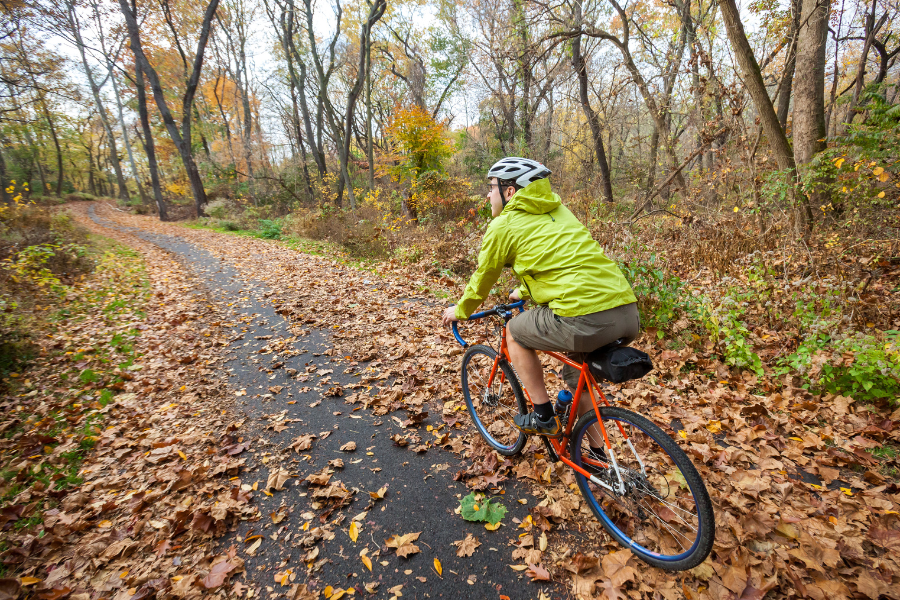 Man in a green rain jacket riding an orange bicycle on a leaf covered trail