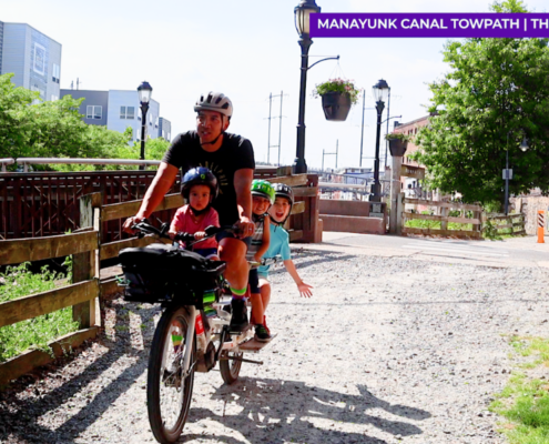 Man riding a cargo bike with three children. One child is on a saddle in the front and two are in a cargo seat in the back.