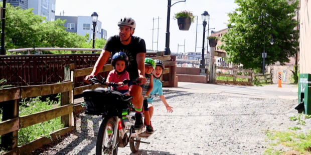 Man riding a cargo bike with three children. One child is on a saddle in the front and two are in a cargo seat in the back.