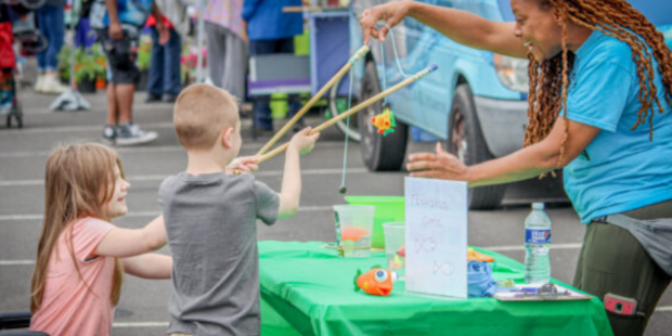 Woman behind a table at an outdoor event showing two children how to use fishing poles.