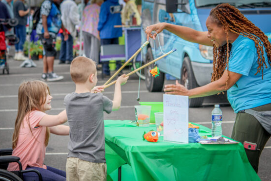 Woman behind a table at an outdoor event showing two children how to use fishing poles.